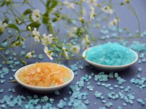 A close-up of colorful bath salts in dishes surrounded by delicate white flowers on a blue surface.